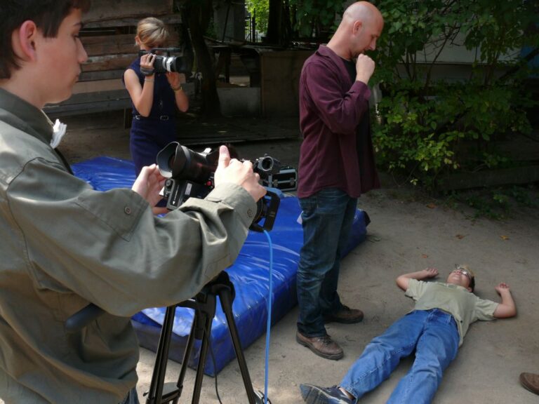 Einblicke in die Filmproduktion: Rechts ein am Boden liegender Junge mit zerbrochener Brille, in der Mitte ein Mann der auf ihn herabschaut, links im Hintergrund ein Mädchen mit einer Videokamera und vorne links ebenfalls ein Junge, der die Videokamera auf die Szene richtet