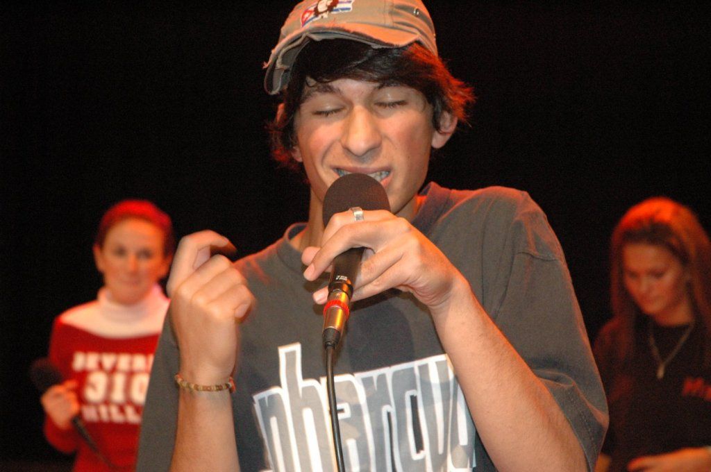 Ein junger Mann in T-Shirt und Cappy, der mit geschlossenen Augen in ein Mikro singt. Links und rechts im Hintergrund zwei junge Frauen, eine von ihnen hat auch ein Mikro.