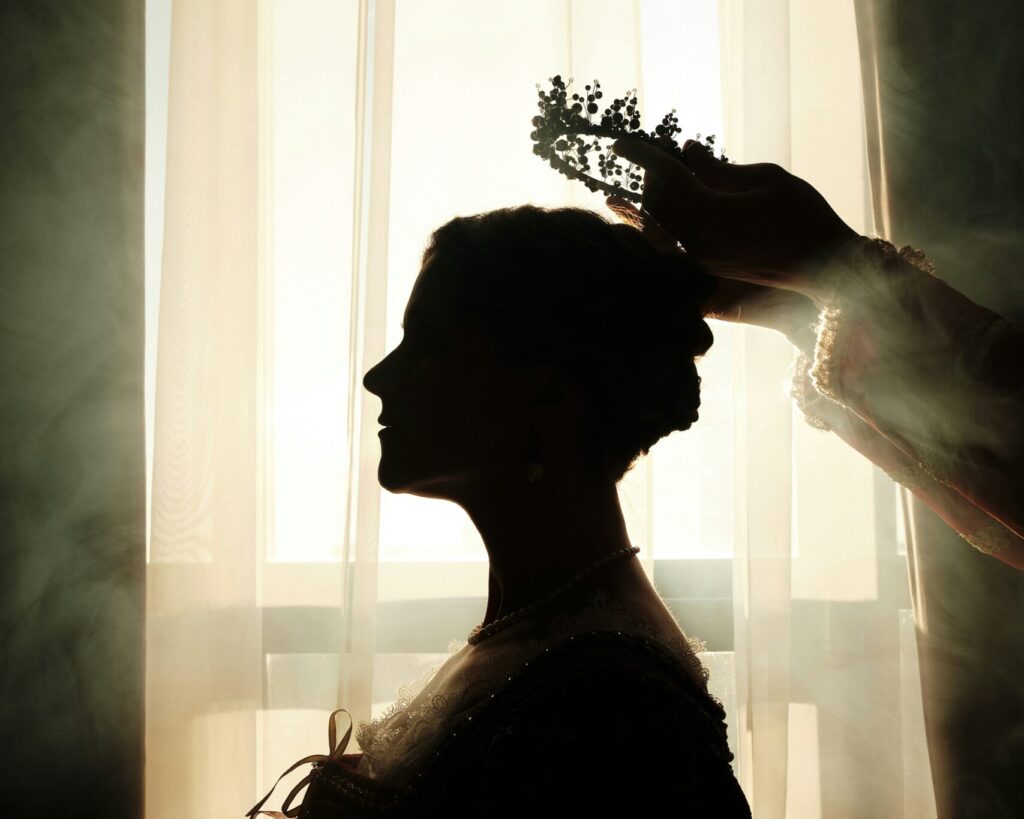 Die Silhouette einer edlen gekleideten Frau vor einem Fenster, durch das die Sonne scheint. Rechts im Bild zwei Arme, die der Frau einen Kranz aufsetzen.