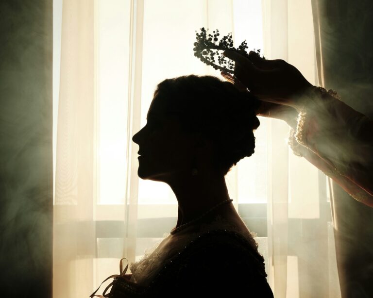 Die Silhouette einer edlen gekleideten Frau vor einem Fenster, durch das die Sonne scheint. Rechts im Bild zwei Arme, die der Frau einen Kranz aufsetzen.