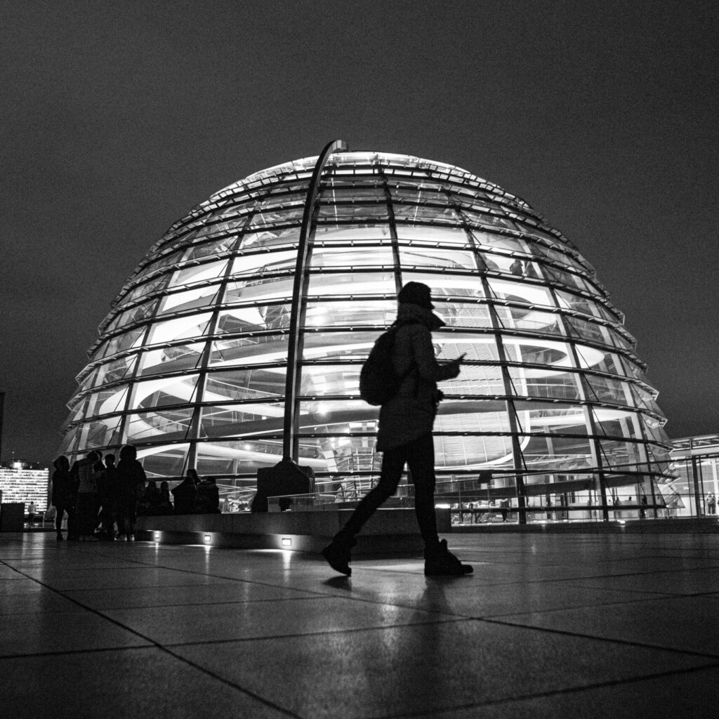 Die Silhouette einer Person vor einer eindrucksvollen Architektur. Die Architektur ist von Innen beleuchtet und spiralförmig aufgebaut, links vor der Architektur ist eine Steinbank platziert auf der mehrere Menschen sitzen.