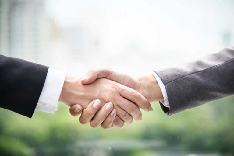 Zwei Arme, die sich in der Mitte des Bildes die Hände schütteln. Beide in formalen Anzügen