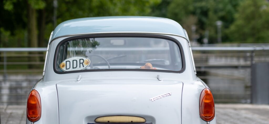 Ein kleines, hellblaues altes Auto von hinten. Im Hintergrund ein Geländer und ein Fluss. An der Heckscheibe des Autos ist ein Schild mit der Aufschrift "DDR" angebracht.