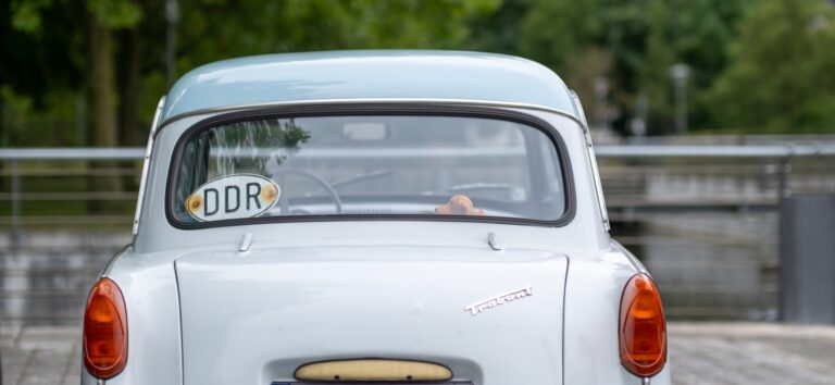 Ein kleines, hellblaues altes Auto von hinten. Im Hintergrund ein Geländer und ein Fluss. An der Heckscheibe des Autos ist ein Schild mit der Aufschrift "DDR" angebracht.