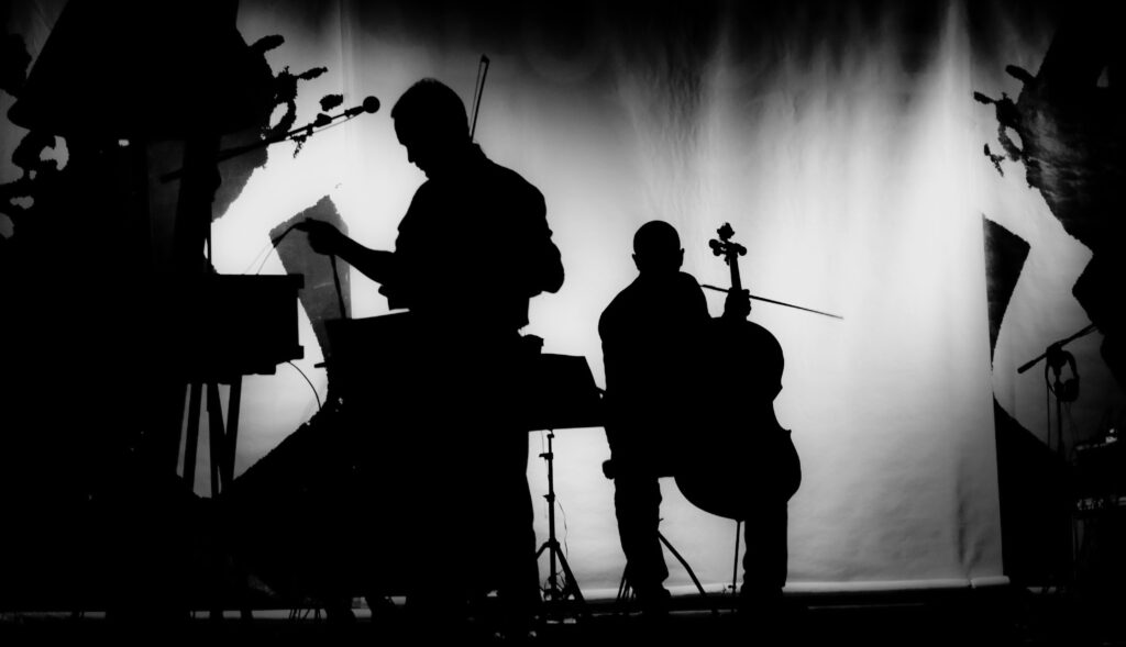 Schwarz-weiß Foto eines Musiksets. In der Mitte sind die Silhouetten zweier Musiker vor einer Leinwand abgebildet, einer davon sitzt an seinem Cello, der andere bereitet sich vermutlich noch vor.