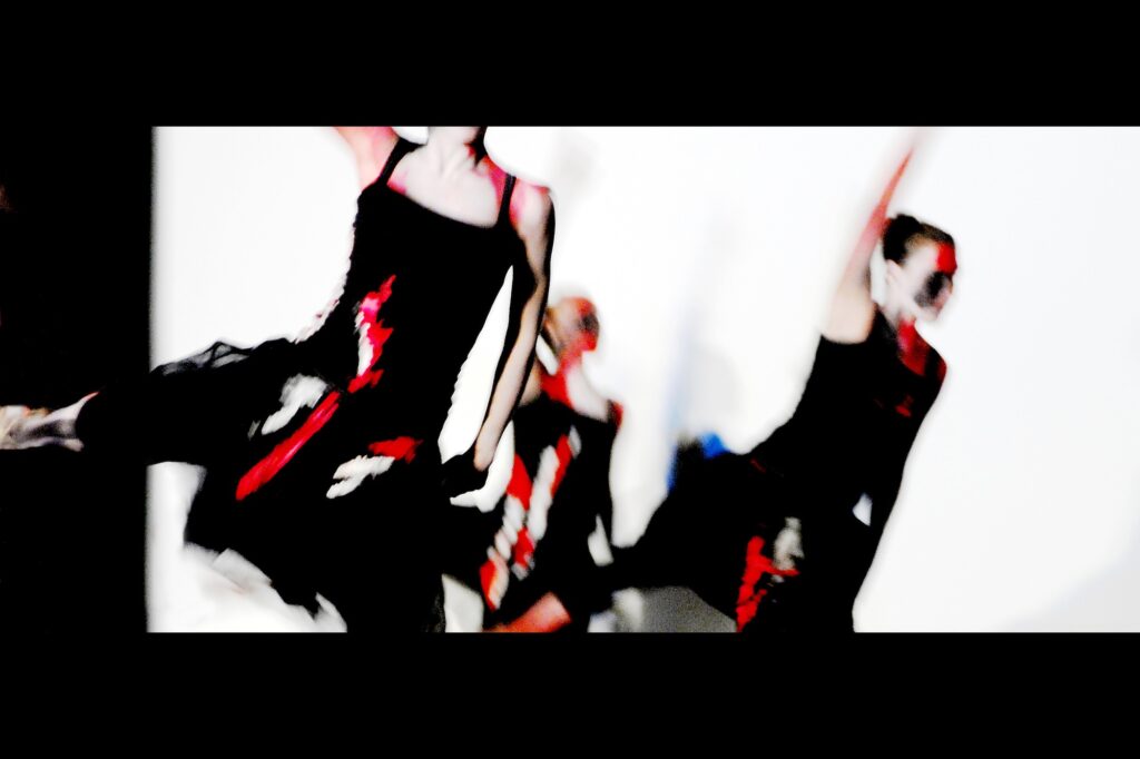 Drei schwarz gekleidete Tänzerinnen in Bewegung. Sie werden von Projektoren vor weißem Hintergrund mit rot-weißen Formen bestrahlt.
