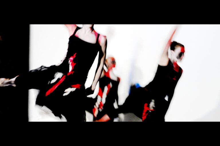 Drei schwarz gekleidete Tänzerinnen in Bewegung. Sie werden von Projektoren vor weißem Hintergrund mit rot-weißen Formen bestrahlt.
