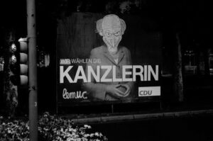 Großes Wahlplakat der CDU am Straßenrand, dass gezielt ins Lächerliche gezogen wird. Über dem Gesicht der vorgeschlagenen Kanzlerin Merkel klebt ein heimtückisches, männliches Gesicht aus einer Cartoon-Serie.