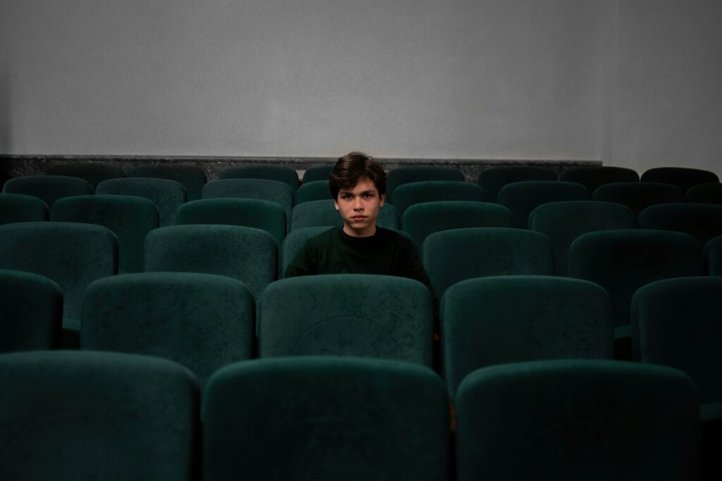 Ein leerer Kinosaal mit dunkelgrünen Sitzen, in der Mitte des Bildes sitzt ein Junge im schwarzen Pullover