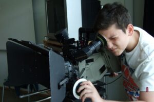 Ein Junge, der konzentriert mit einem Auge durch eine große, schwarze Filmkamera blickt