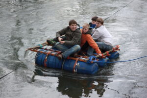 Vier Jungs auf einem aus Fässern gebauten Floß. Der junge vorne am Rande des Floß, zieht dieses entlang einer Leine während sein Hintermann ängstlich aussieht.