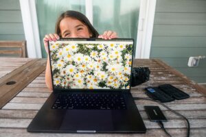 Ein fröhliches junges Mädchen, dass an einem Holztisch sitzt und von hinten über den Bildschirm eines Laptops schaut. Der Bildschirm zeigt ein Feld weißer Blumen. Rechts neben dem Laptop liegt eine Kamera.