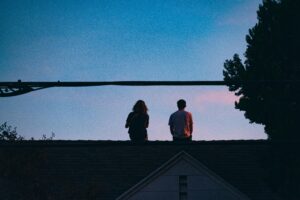 Zwei junge Menschen von hinten, die auf einem Ziegeldach sitzen im Licht der Abenddämmerung. Quer durch das Bild zieht sich die Oberleitung, rechts befindet sich ein Baum.