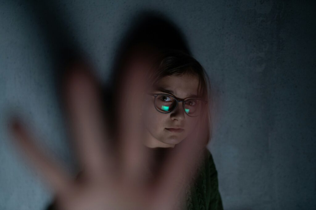 Eine ängstlich schauende, junge Person mit Brille vor einer grauen Wand, die ihre Hand vor die Linse hält.