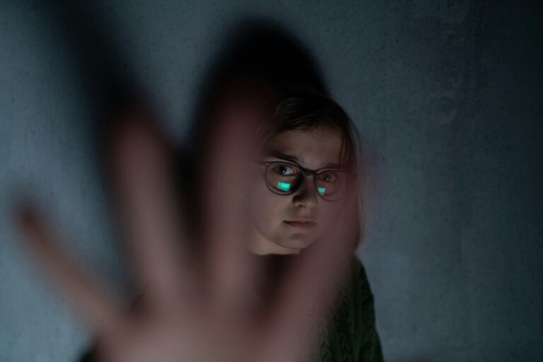 Eine ängstlich schauende, junge Person mit Brille vor einer grauen Wand, die ihre Hand vor die Linse hält.