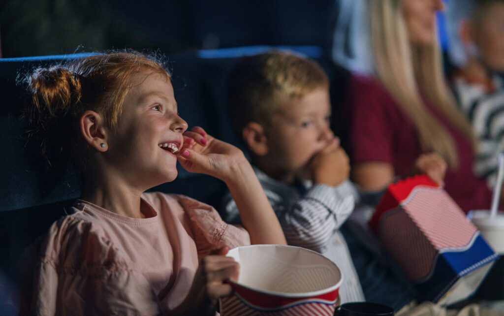 Zwei Kinder sitzen im Kino, essen Popcorn und schauen einen Film mit viel Freude im Gesicht.