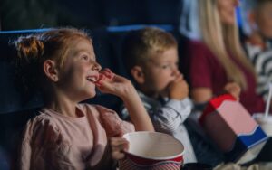 Zwei Kinder sitzen im Kino, essen Popcorn und schauen einen Film mit viel Freude im Gesicht.