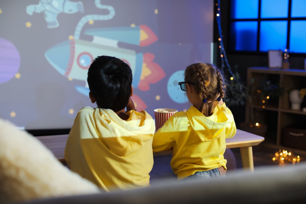 Zwei Kinder schauen auf einer großen Leinwand Zuhause einen Kinderfilm. Es ist ein Weltall mit großer Rakete zu sehen.