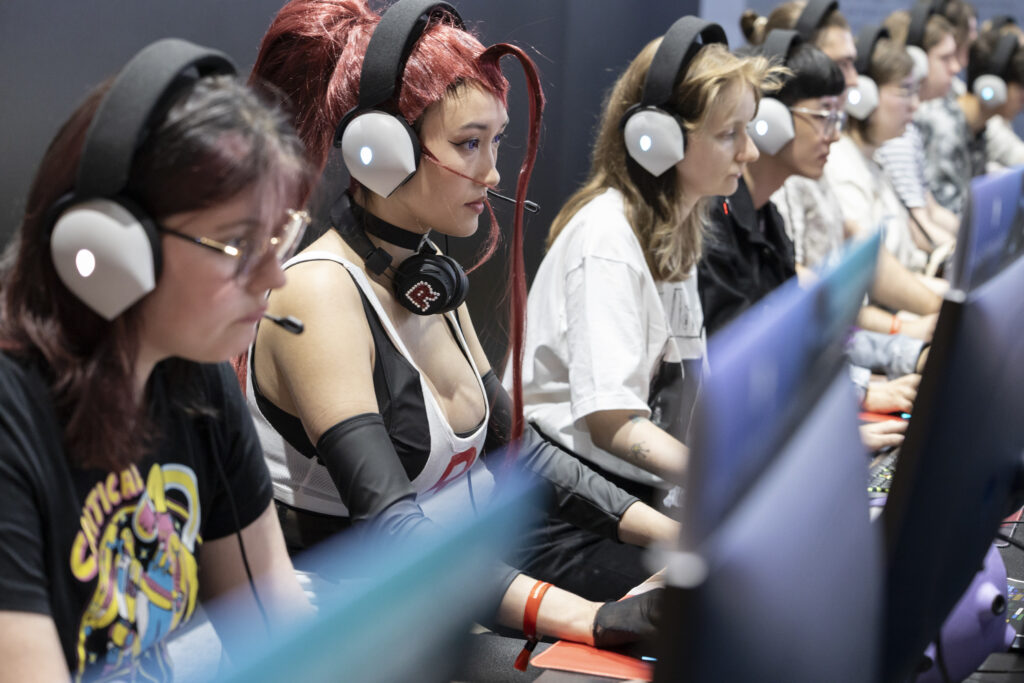 Mehrere Frauen mit Headset, eine in Cosplay-Outift, sitzen an Computern auf der Gamescom und spielen Computerpiele