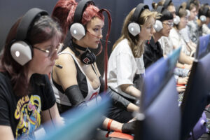 Mehrere Frauen mit Headset, eine in Cosplay-Outift, sitzen an Computern auf der Gamescom und spielen Computerpiele
