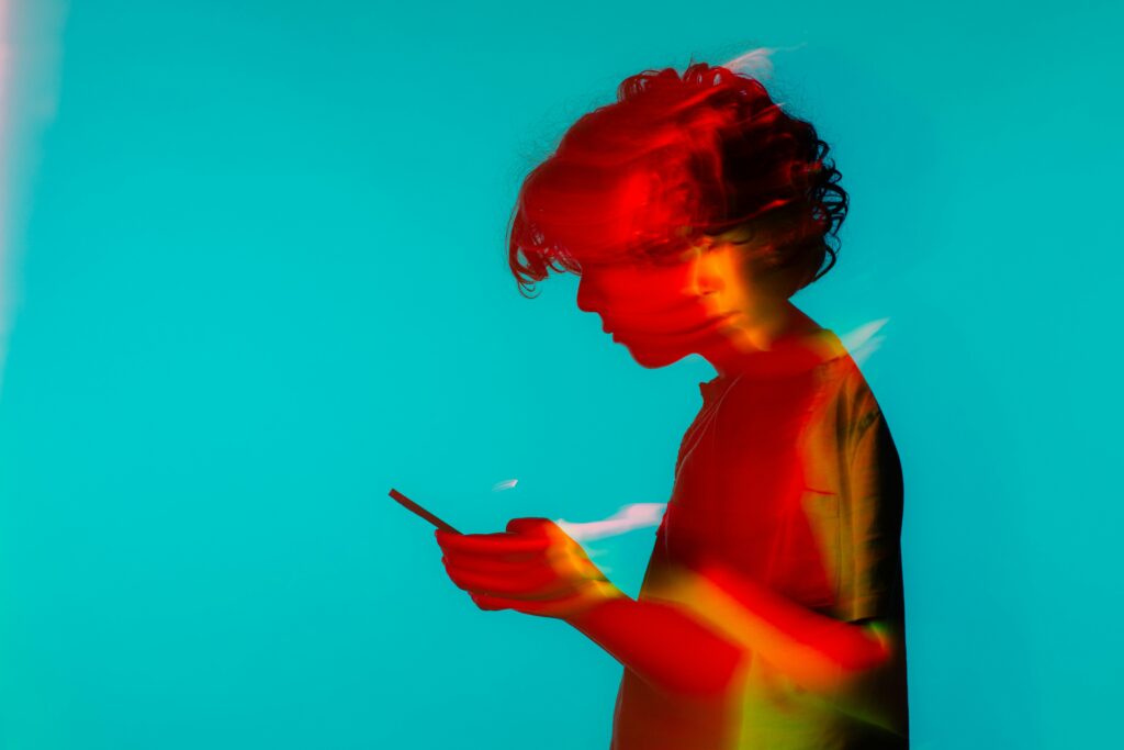 Ein Jugendlicher hält ein Smartphone in den Händen. Er steht vor blauem Hintergrund, sein Körper ist digital in rot/gelb eingefärbt und sein Kopf verschwommen