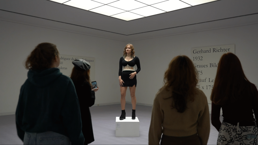 In der Mitte steht eine junge Frau im schwarzen Kleid auf einem Sockel, wie ein Ausstellungsstück. Im Vordergrund sind mehrere Besuchende zu sehen, die die Frau betrachten. Links und Rechts an den Wänden des Ausstellungsraumes sind große Tafeln montiert.