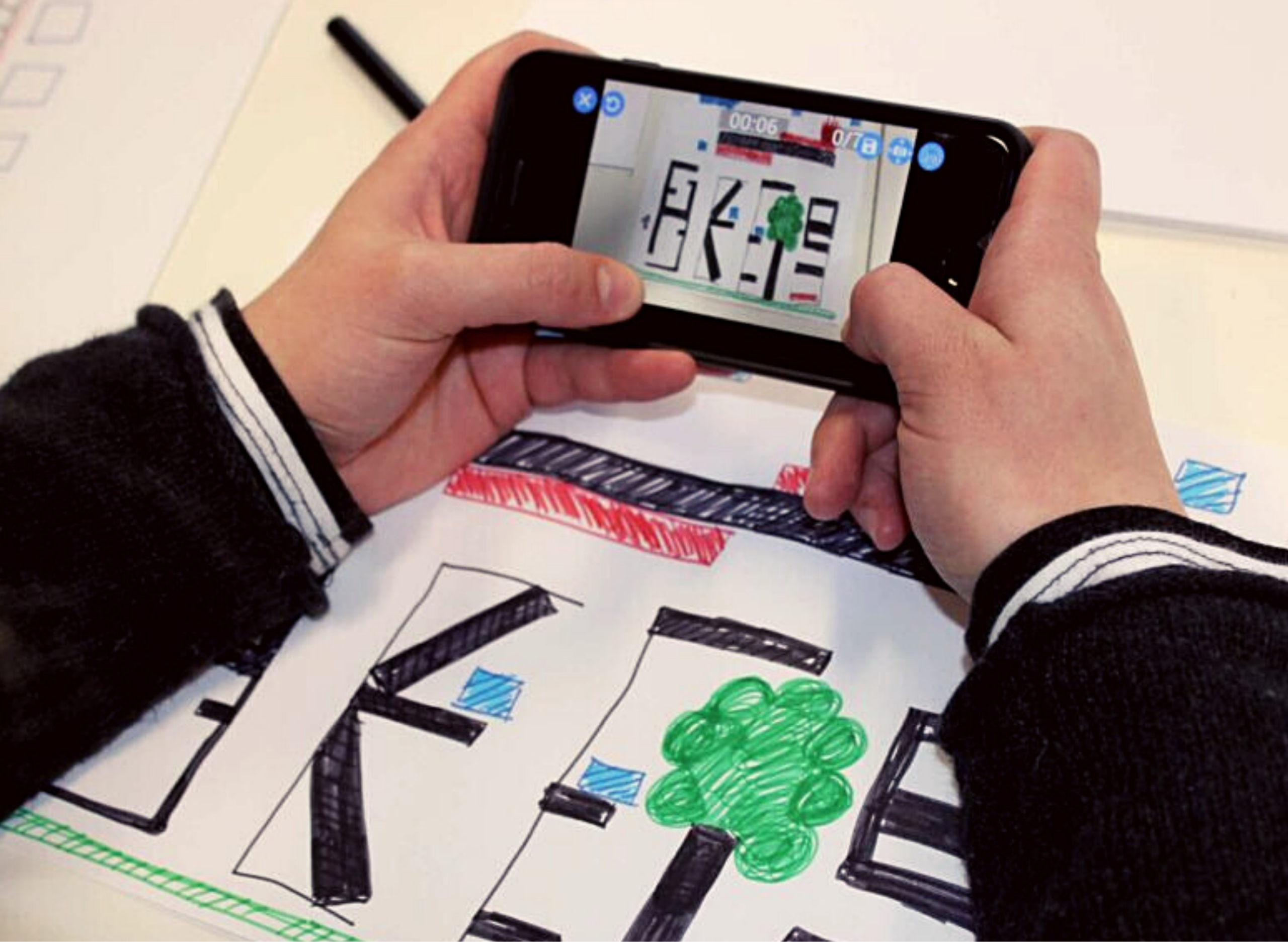 Hände eines Kindes, welches ein Smartphone darin Hält. Abgestützt auf einem gemalten Bild. Auf dem Handy sieht man das selbe Bild, in der App "Draw your Game".