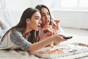 Zwei junge Frauen liegen auf dem Boden, sehen fern und essen dabei Pizza