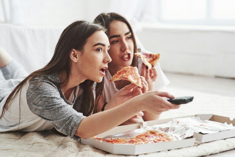 Zwei junge Frauen liegen auf dem Boden, sehen fern und essen dabei Pizza