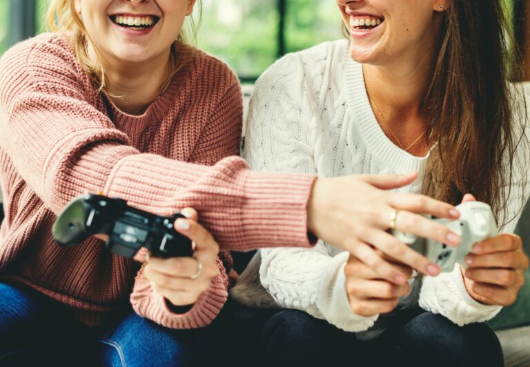 Zwei Frauen spielen lachend zusammen Playstation und halten dafür Controller in den Händen.
