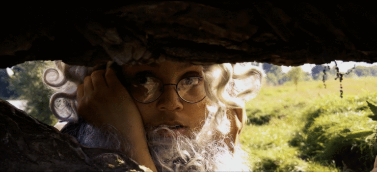 Verkleidetes Kind schaut durch einen Felsspalt in grüner Landschaft.