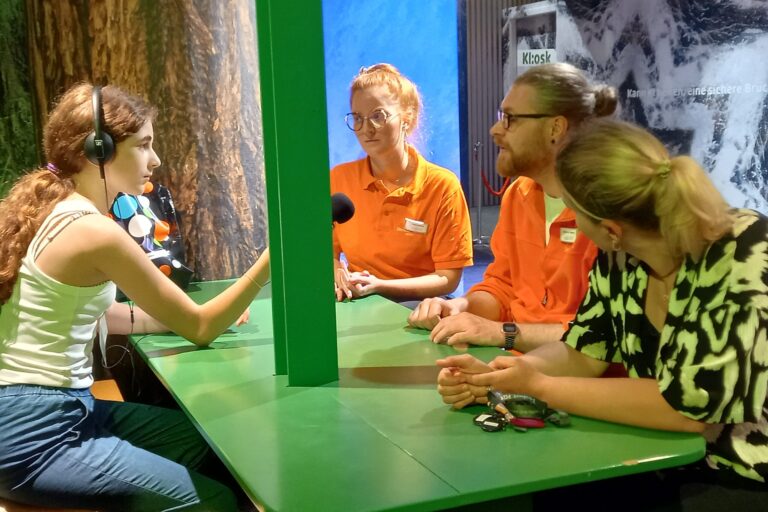 Mädchen interviewt drei Personen. Sie sitzen an einem grünen Tisch.