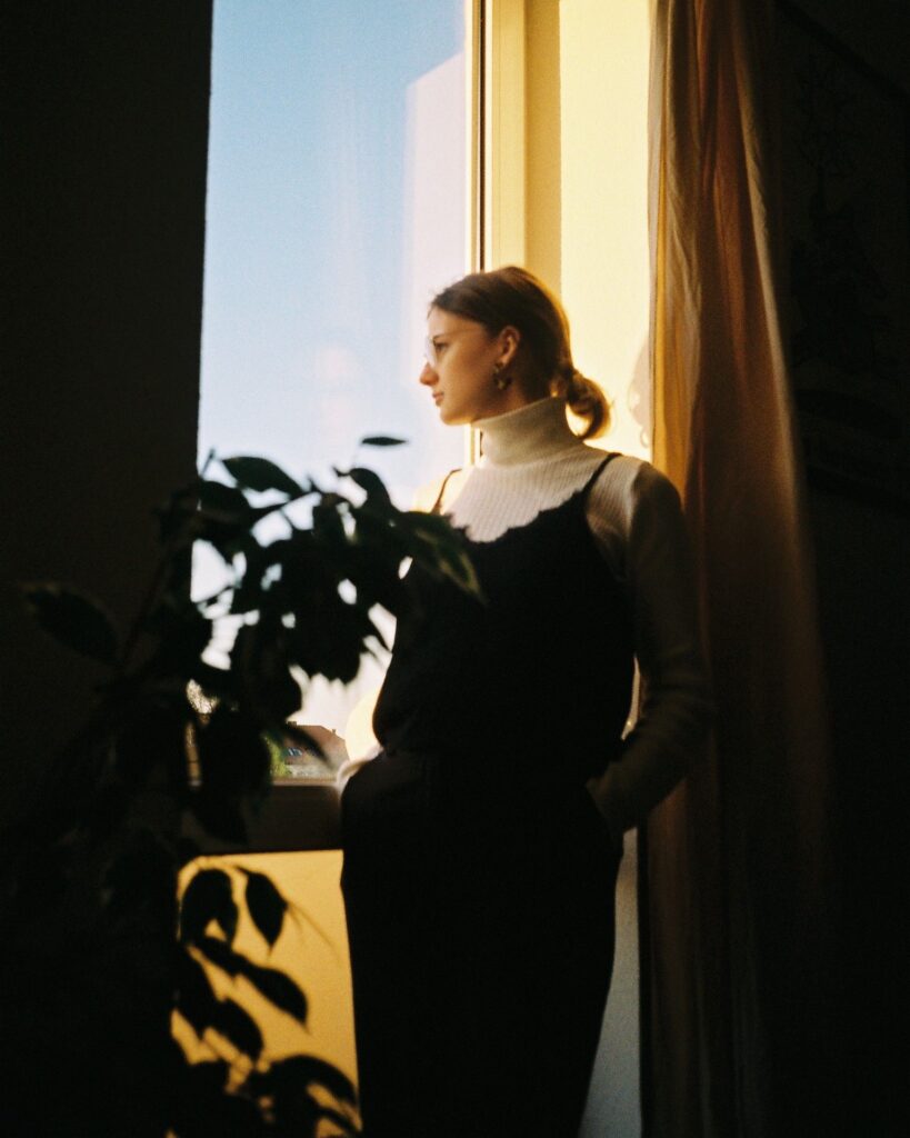 Eine Frau die ihre Hände in den Hosentaschen hält, steht vor einem Fenster und schaut nachdenklich hinaus. Das Fenster ist die einzige Lichtquelle, sodass der Rest des Bildes Schatten sind. Im Vordergrund ist der Schatten einer Pflanze zu sehen.