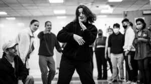 Schwarz-weiß Bild auf dem eine Ansammlung von Menschen um eine tanzende Frau steht/sitzt.