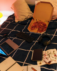 Ein Bett auf dem eine angefangene Pizza in der Schachtel, ein Laptop und Schreibzeug liegt.