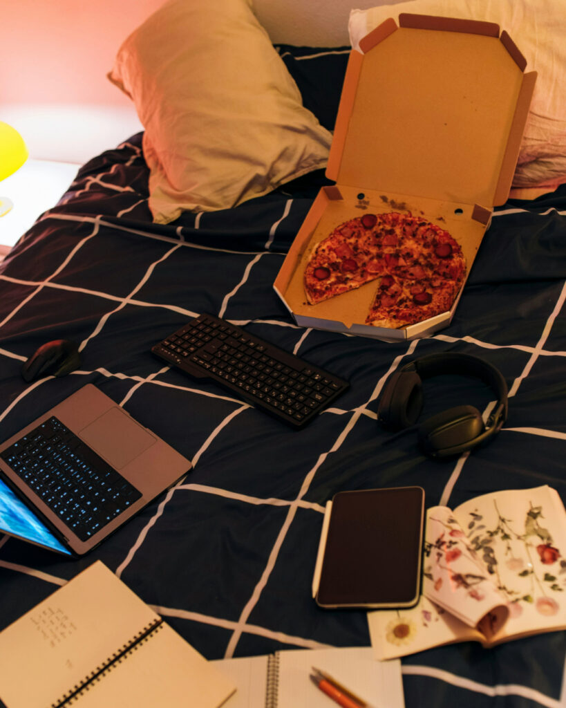 Ein Bett auf dem eine angefangene Pizza in der Schachtel, ein Laptop und Schreibzeug liegt.