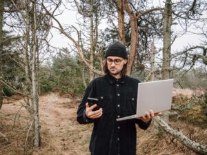 Mann steht mitten im Wald (Dünen) mit Laptop und Smartphone in der Hand.