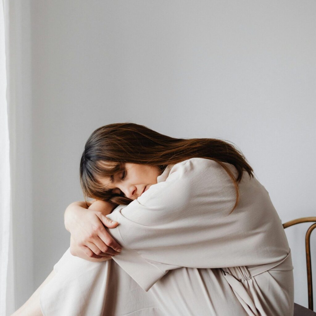 Frau in weißer Kleidung, auf einem Tisch sitzend, die ihren Kopf auf ihre Arme abgelegt hat
