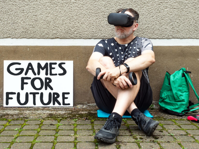 Ein Mann der mit aufgesetzter VR-Brille und Controllern draußen auf dem Boden sitzt, links neben ihm ein Schild mit der Aufschrift "Games for Future"