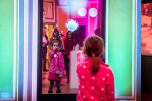 Ein Mädchen steht vor einem Spiegel/einer digitalen Installation und versucht mit ihrem Arm Icons zu bewegen