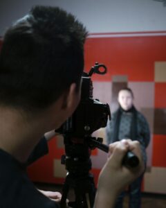 Eine junge Person, die durch eine Filmkamera schaut und diese steuert, während im Hintergrund eine junge Frau vor einer bunten Wand steht