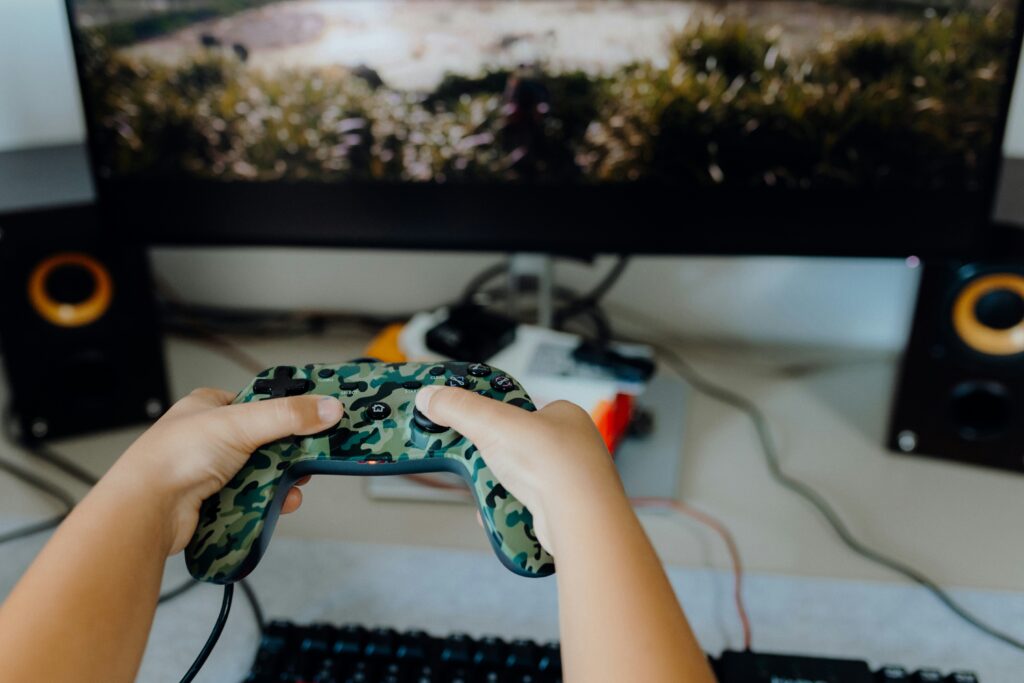 Hände halten einen Controller für eine Konsole in Camouflage-Optik in den Händen, im Hintergrund sieht man einen großen Bildschirm und Computerboxen.