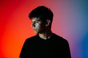 Ein junger Mann in schwarzem Shirt steht vor einer bunten Wand mit Farbverlauf von rot zu lila zu blau