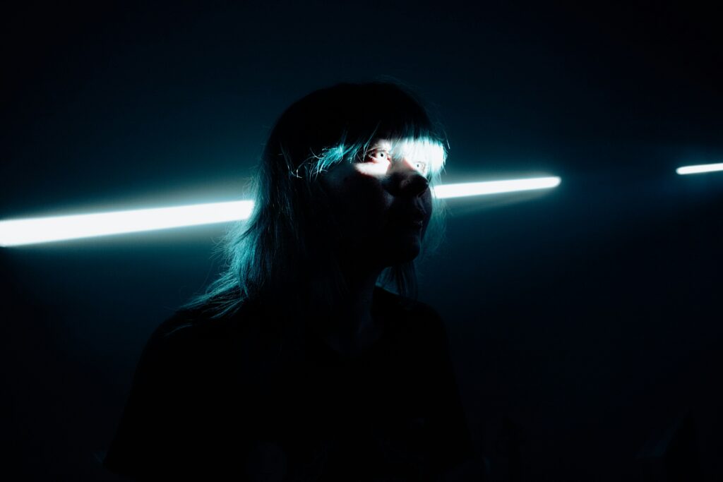 Eine junge Frau mit blauen Haaren in einem dunklen Raum. Durch einen Spalt fällt Licht, das ihre Augen beleuchtet.