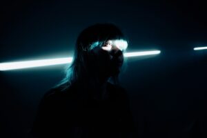 Eine junge Frau mit blauen Haaren in einem dunklen Raum. Durch einen Spalt fällt Licht, das ihre Augen beleuchtet.