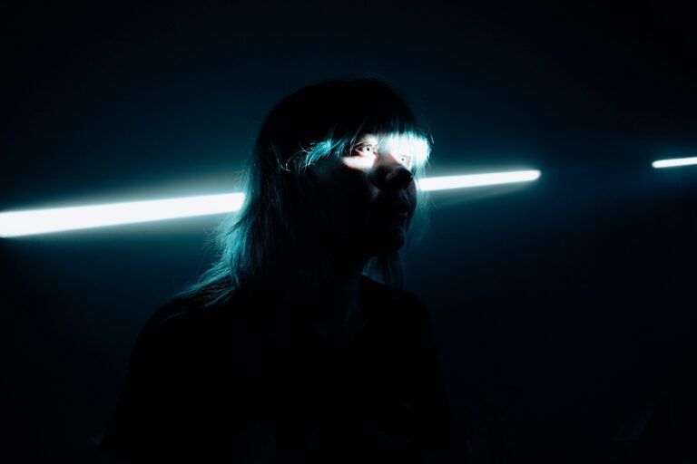 Eine junge Frau mit blauen Haaren in einem dunklen Raum. Durch einen Spalt fällt Licht, das ihre Augen beleuchtet.