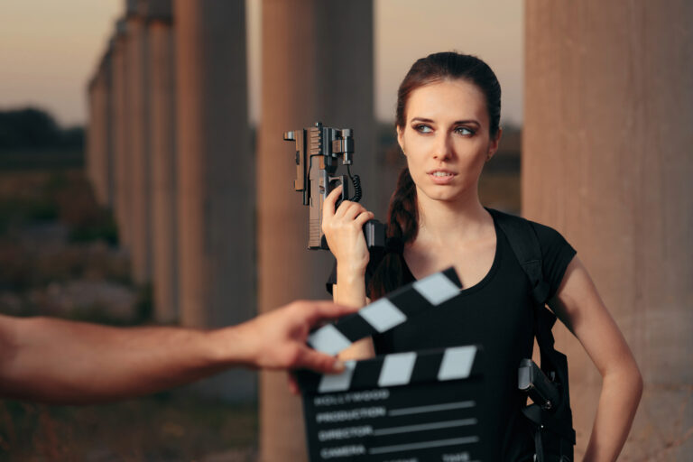 Eine Frau steht in einem Spielfilmset mit einer Waffe in der Hand. Im Vordergrund in der Unschärfe wird eine Filmklappe geschlagen.