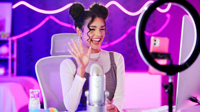 Eine jugendliche Influencerin (gestellt/Model/unbekannt) sitzt vor einem Ringlicht und vor einem Mikrofon und strahlt und winkt in die Kamera. Ihr Zimmer ist pink beleuchtet