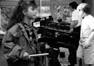 Ein junges Mädchen, dass eine große, professionelle Filmkamera bedient. Rechts im Hintergrund zwei weitere Personen.