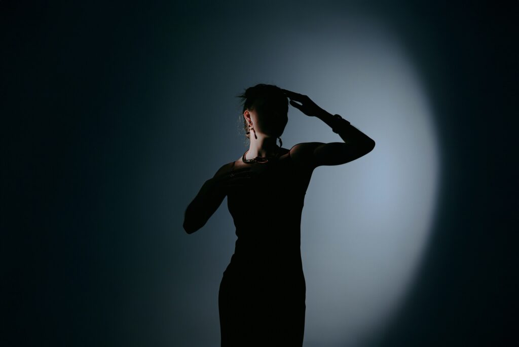 schwarze Silhouette einer jungen Frau, die vor dunklem Hintergrund posiert. Nur am Hals wird die Frau von einem Licht angestrahlt.